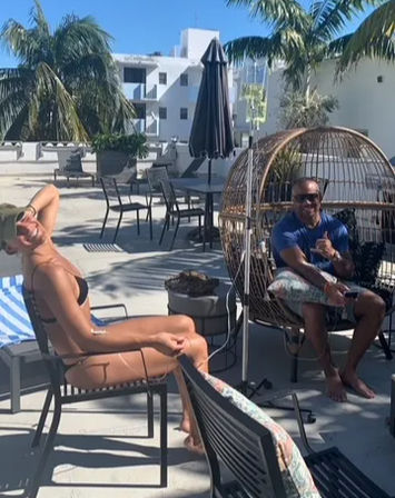 Sunny rooftop pool deck with palm trees; woman in a black bikini lounges on a chair while a man in sunglasses flashes a shaka from a wicker egg chair, patio umbrellas and modern white buildings in the background.