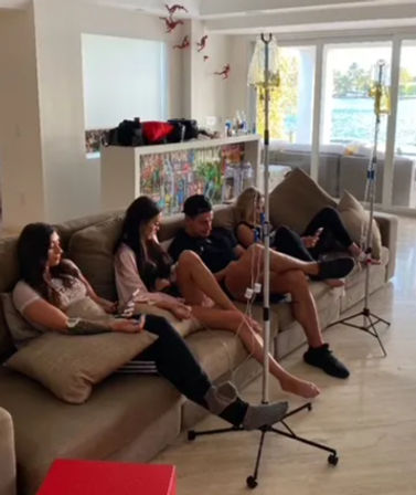 Four people lounging on a sectional sofa receiving IV hydration drips from poles in a bright waterfront living room with large windows.