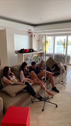 Group of adults relaxing on a beige sectional couch in a bright modern living room receiving IV drips from tall infusion stands, with floor-to-ceiling glass doors revealing a sunny waterfront view.