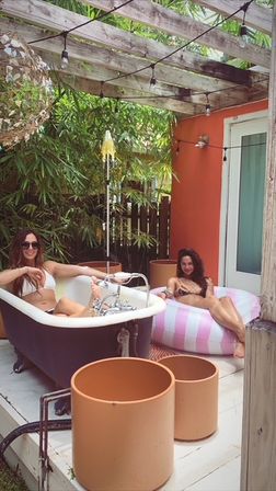 Backyard patio scene with two women in bikinis relaxing on a pink-striped inflatable lounger and a freestanding black-and-white bathtub under a wooden pergola with string lights, bamboo privacy greenery and terracotta planters.