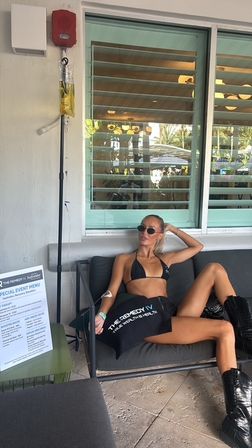 Woman in black bikini and boots lounging on an outdoor patio sofa under a shuttered window with palm-tree reflections, wearing sunglasses and reclining beside an IV drip pole — sunny poolside relaxation.