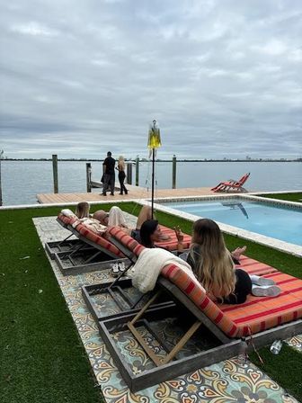 Relaxing waterfront pool patio with striped chaise lounges on patterned tile and turf, rectangular pool and wooden dock on a calm bay under an overcast sky.