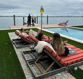 Several people relaxing on striped red-orange chaise lounges beside a rectangular pool and artificial turf on a waterfront deck, with a wooden dock and two walkers by calm water under an overcast sky.