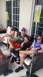 Three men lounging on a shaded balcony patio with IV hydration bags on stands; casual summer attire, one wearing a purple novelty crown, another in sunglasses, drinks and sandals scattered nearby.