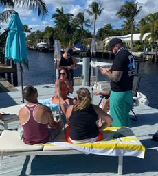 Five people socializing on a sunny waterfront dock by a canal with palm trees, lounge chairs, a turquoise umbrella and a boat in the background.
