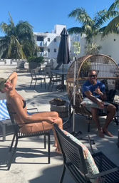 Sunny rooftop pool deck with palm trees, lounge chairs and umbrellas; a woman in a black bikini reclines while a man in sunglasses relaxes in a woven egg chair flashing a shaka — laid-back tropical vacation vibe.