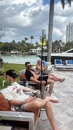 Three men relaxing on outdoor lounge chairs at a tropical waterfront marina with palm trees and a luxury yacht nearby, two IV hydration bags on stands visible under a cloudy sky.