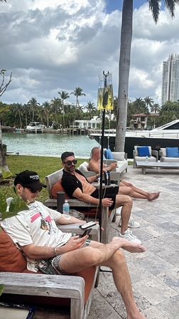 Three men relaxing on outdoor lounge chairs at a tropical waterfront marina with palm trees and a luxury yacht nearby, two IV hydration bags on stands visible under a cloudy sky.