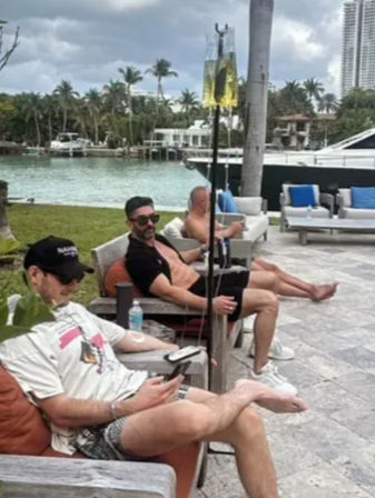 Three men lounging on cushioned patio chairs at a waterfront marina with palm trees and a docked yacht in the background under a cloudy sky.