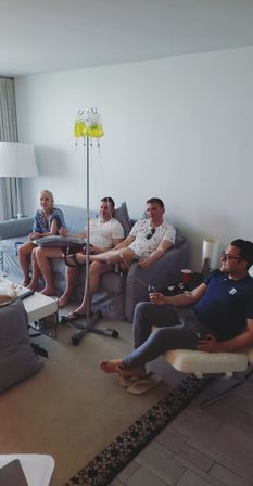 Four adults lounging in a bright modern living room receiving yellow vitamin IV drips from a central pole — relaxed at-home wellness session on couches and chair.
