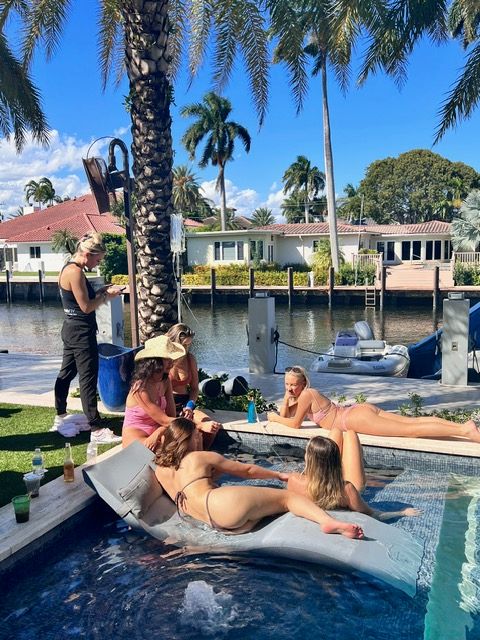 Sunny canal-front backyard with palm trees: a group of people in swimsuits lounging on an inflatable float and pool edge next to a docked dinghy and waterfront homes across the canal.