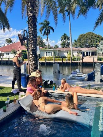 Sunny canal-front backyard with palm trees: a group of people in swimsuits lounging on an inflatable float and pool edge next to a docked dinghy and waterfront homes across the canal.