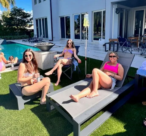 Three friends in colorful swimsuits lounging poolside at a luxury modern home's backyard patio with gray loungers, artificial turf and a turquoise pool on a sunny day