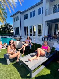 Three friends in bikinis and sunglasses lounging on sunbeds by a turquoise pool at a modern two-story home with palm trees and a bright blue sky