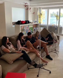 Group IV therapy at home: four people reclined on a beige sofa hooked to IV poles in a bright waterfront living room with floor-to-ceiling windows and water view.