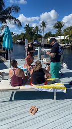 Friends lounging on a sunny waterfront dock beside a palm-lined canal, with lounge chairs, a turquoise umbrella and a boat in the background.