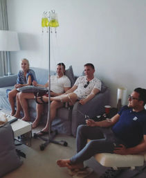 Four adults relaxing on sofas in a bright living room, each receiving yellow vitamin IV drips from a central stand — at-home group IV therapy session.