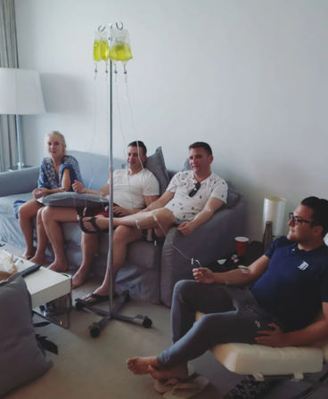 Four adults relaxing on sofas in a bright living room, each receiving yellow vitamin IV drips from a central stand — at-home group IV therapy session.