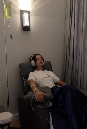 Person relaxing in an IV therapy lounge chair with headphones on, receiving an IV drip through their arm and covered by a navy blanket under soft wall lighting.