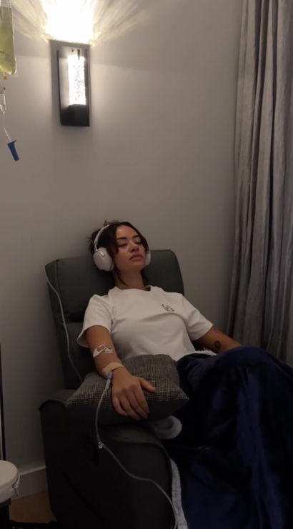 Person relaxing in an IV therapy lounge chair with headphones on, receiving an IV drip through their arm and covered by a navy blanket under soft wall lighting.