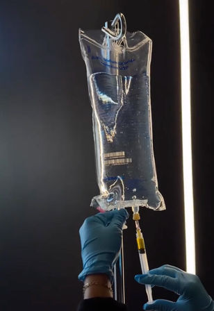 Close-up of hands in blue medical gloves withdrawing clear saline from a hanging IV fluid bag with a syringe, backlit against a dark hospital or clinic background.