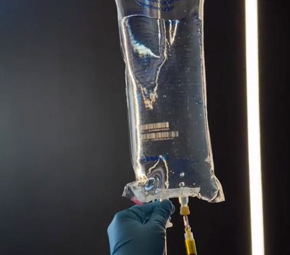 Close-up of hands in blue medical gloves withdrawing clear saline from a hanging IV fluid bag with a syringe, backlit against a dark hospital or clinic background.