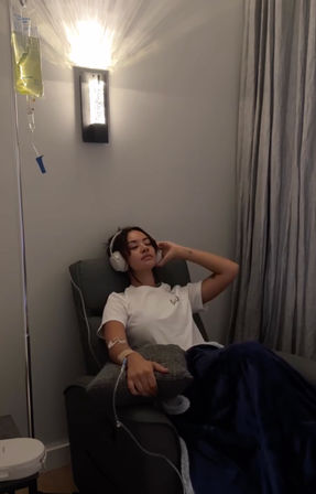 Person reclining in an infusion chair wearing headphones and a white T‑shirt, receiving IV therapy from a hanging drip, covered with a navy blanket under a soft wall light and gray curtains.
