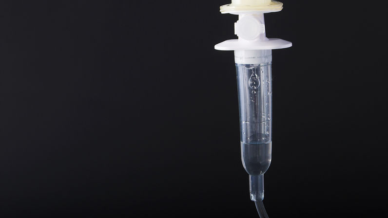 Close-up of a clear intravenous (IV) drip chamber with saline and rising air bubbles, tubing hanging against a dark background