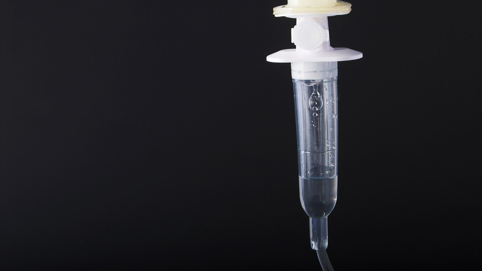 Close-up of a clear intravenous (IV) drip chamber with saline and rising air bubbles, tubing hanging against a dark background