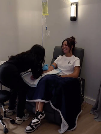 Smiling woman in a wellness clinic recliner receiving an IV drip as a technician adjusts the tubing, navy blanket over her legs.