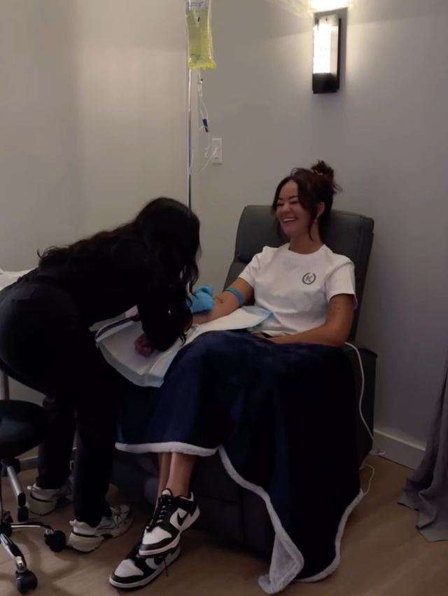 Smiling woman in a wellness clinic recliner receiving an IV drip as a technician adjusts the tubing, navy blanket over her legs.