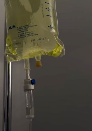 Yellow-tinted IV drip bag hanging from a chrome IV pole with a clear drip chamber and tubing visible against a gray clinical wall.