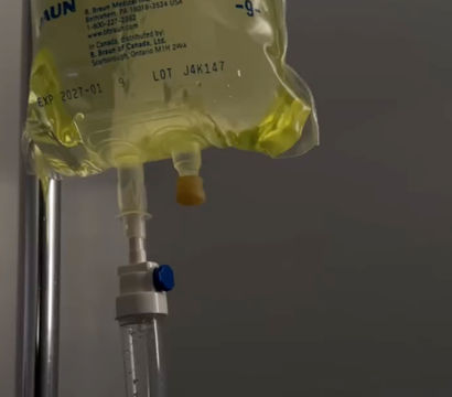 Yellow-tinted IV drip bag hanging from a chrome IV pole with a clear drip chamber and tubing visible against a gray clinical wall.