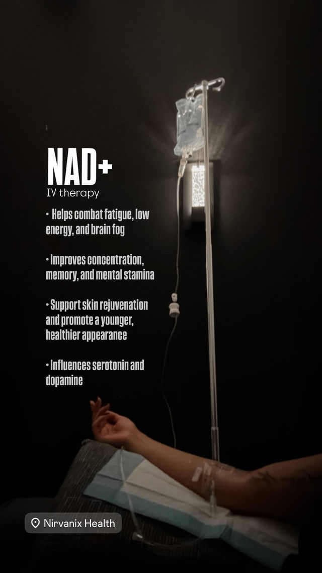 Dimly lit wellness clinic room showing a NAD+ IV therapy bag on a pole and a forearm with an IV catheter resting on a pillow, with promotional bullet points about energy, focus and skin rejuvenation visible on the wall.
