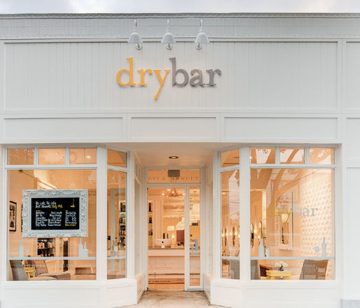 Chic white blowout hair salon storefront with large display windows, warm interior lighting, framed chalkboard menu and inviting central entrance.