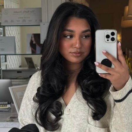 Mirror selfie of a person with long dark glossy waves and neutral makeup wearing a cream cardigan, holding a white iPhone in a hair salon setting.