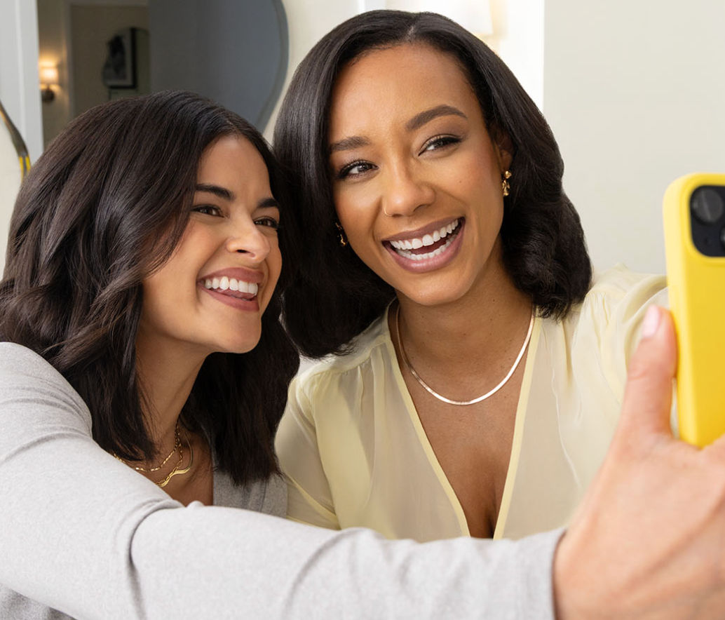 Two friends laughing and taking a close-up indoor selfie with a yellow phone, bright casual portrait.