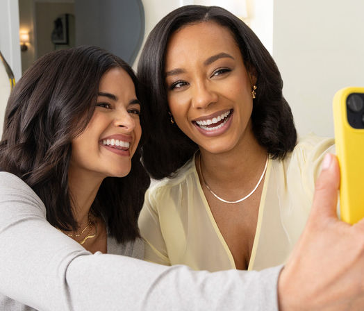 Two friends laughing and taking a close-up indoor selfie with a yellow phone, bright casual portrait.