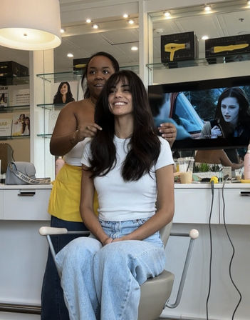 Smiling woman with long dark hair sits in a salon chair wearing a white tee and light jeans as a stylist adjusts her layers in a bright modern hair salon.