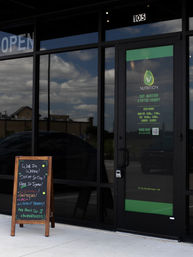 Glass-front wellness clinic storefront with glowing OPEN sign, green IV therapy poster on the door, chalkboard sidewalk sign advertising walk-ins, and cloudy sky reflections in the glass.
