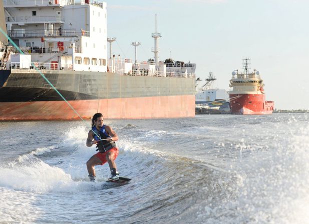 Wakeboarder in red shorts and life vest carving through a wake near a large cargo ship and red supply vessel in a busy harbor, water spray glinting in late afternoon light.