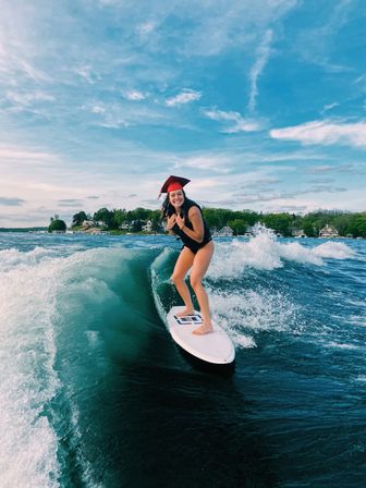 Cheerful woman wearing a red graduation cap wake-surfing on a white board across a blue lake, passing waterfront cottages beneath a bright, cloud-streaked sky.