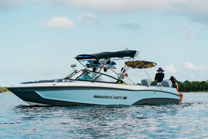 Light-blue wakeboard boat with a black bimini and wakeboard mounted on its tower, people relaxing on board during a sunny, partly cloudy lake day with a tree-lined shore