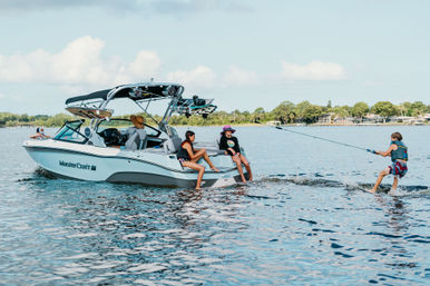 Friends on a white wakeboat on a sunny lake; two sitting on the stern while a life-vested rider is pulled on a wakeboard across sparkling blue water with a tree-lined shoreline.