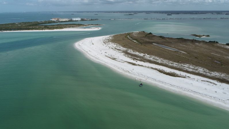 Aerial drone view of a curved white‑sand barrier island with turquoise coastal waters and a small boat along the shoreline.