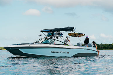 Light-blue wakeboard boat with a black bimini and wakeboard mounted on its tower, people relaxing on board during a sunny, partly cloudy lake day with a tree-lined shore