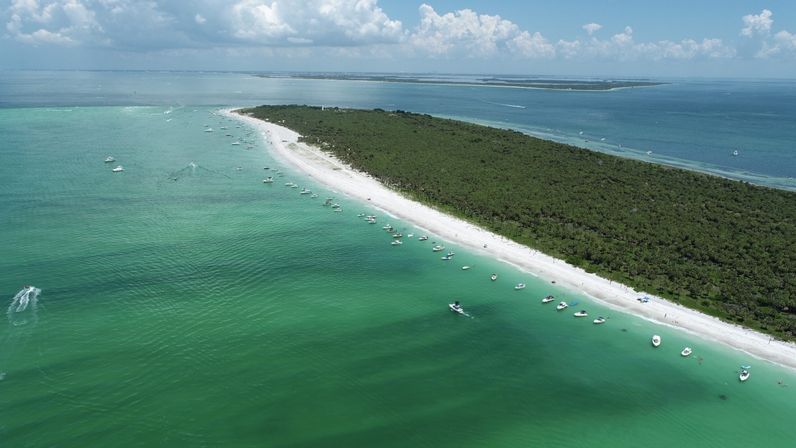 Aerial view of a curved barrier island with dense green trees, white-sand beach and turquoise coastal water dotted with anchored boats under a partly cloudy sky.