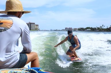 Wakesurfer carving a boat wake near coastal high-rise condos and palm trees on a sunny day