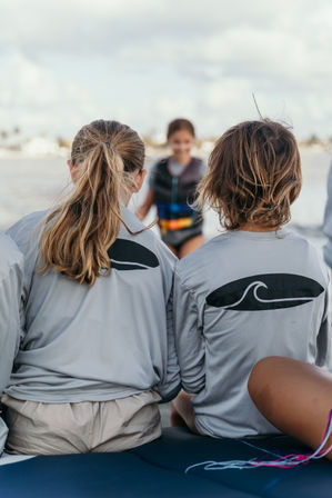 Two kids in matching gray rash guards with surfboard wave logos sit on a boat watching a friend in a colorful life jacket on coastal waters under a cloudy sky