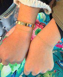 Close-up of two wrists wearing delicate gold chain bracelets and a gold link charm bracelet with a blue evil-eye tile, set against a bright floral fabric background.